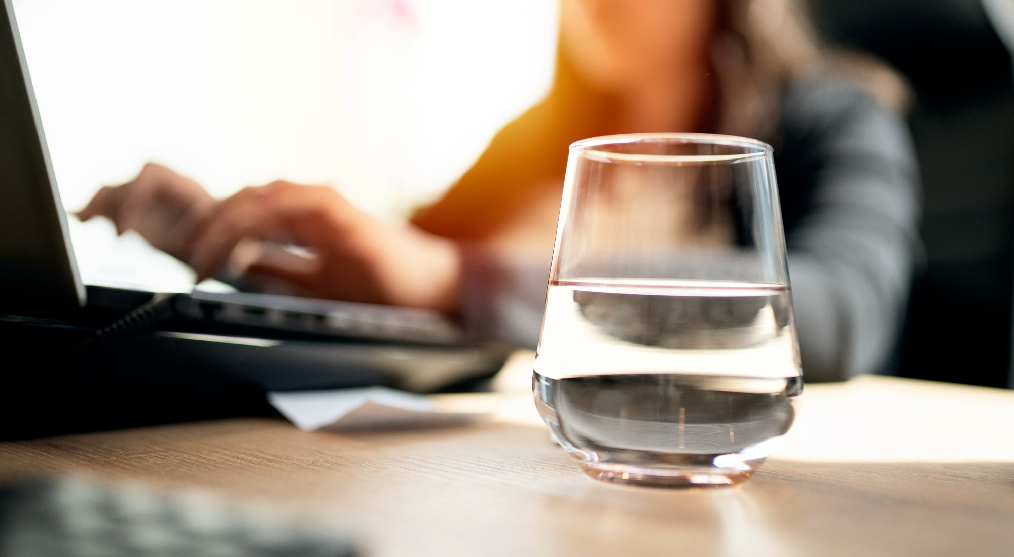 A person is working on their laptop in the background, and in the foreground there is a fancy glass with clear, pure water in it.