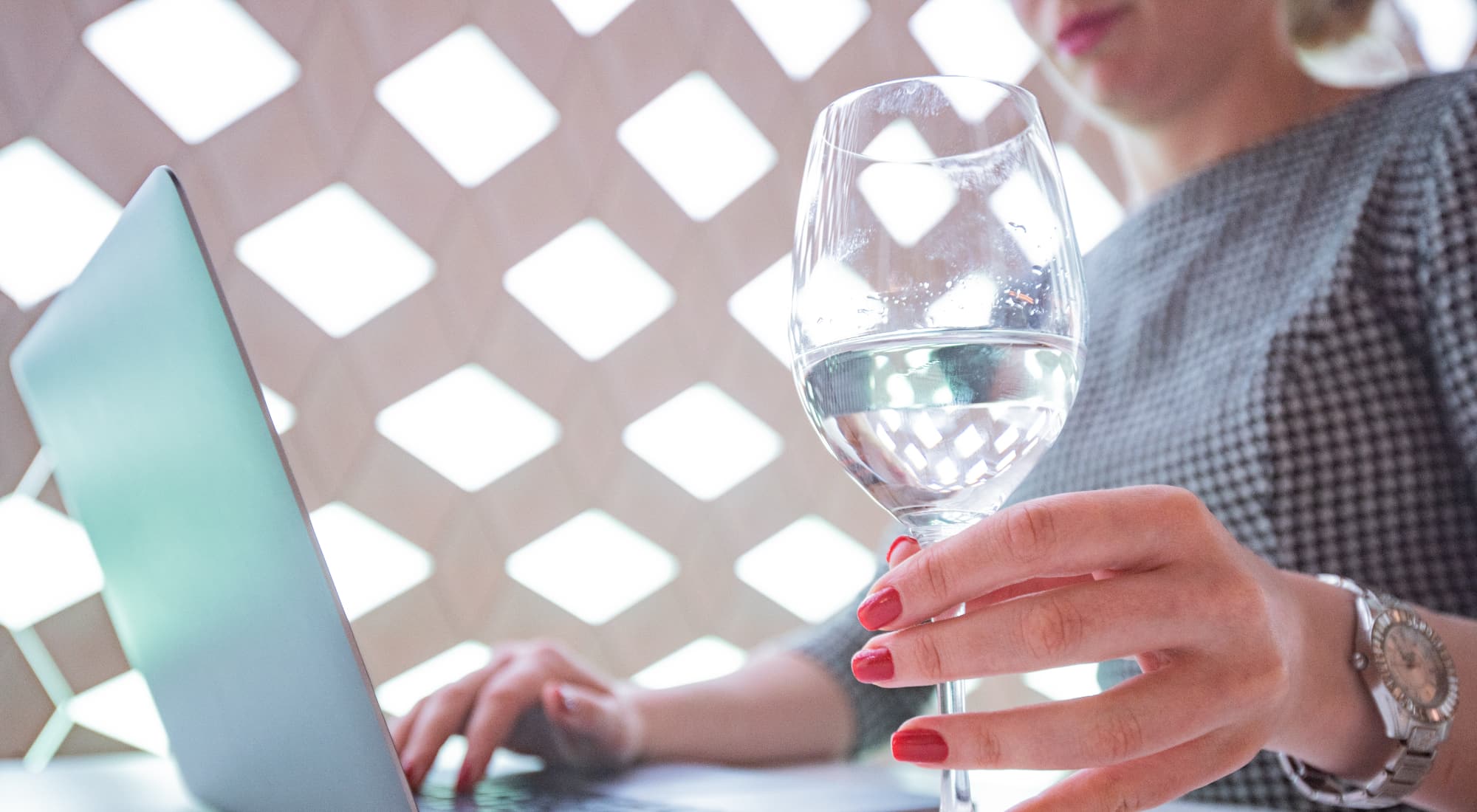 A woman is working on her laptop, and she is holding a wine glass in her hand that has clear, pure water in it.