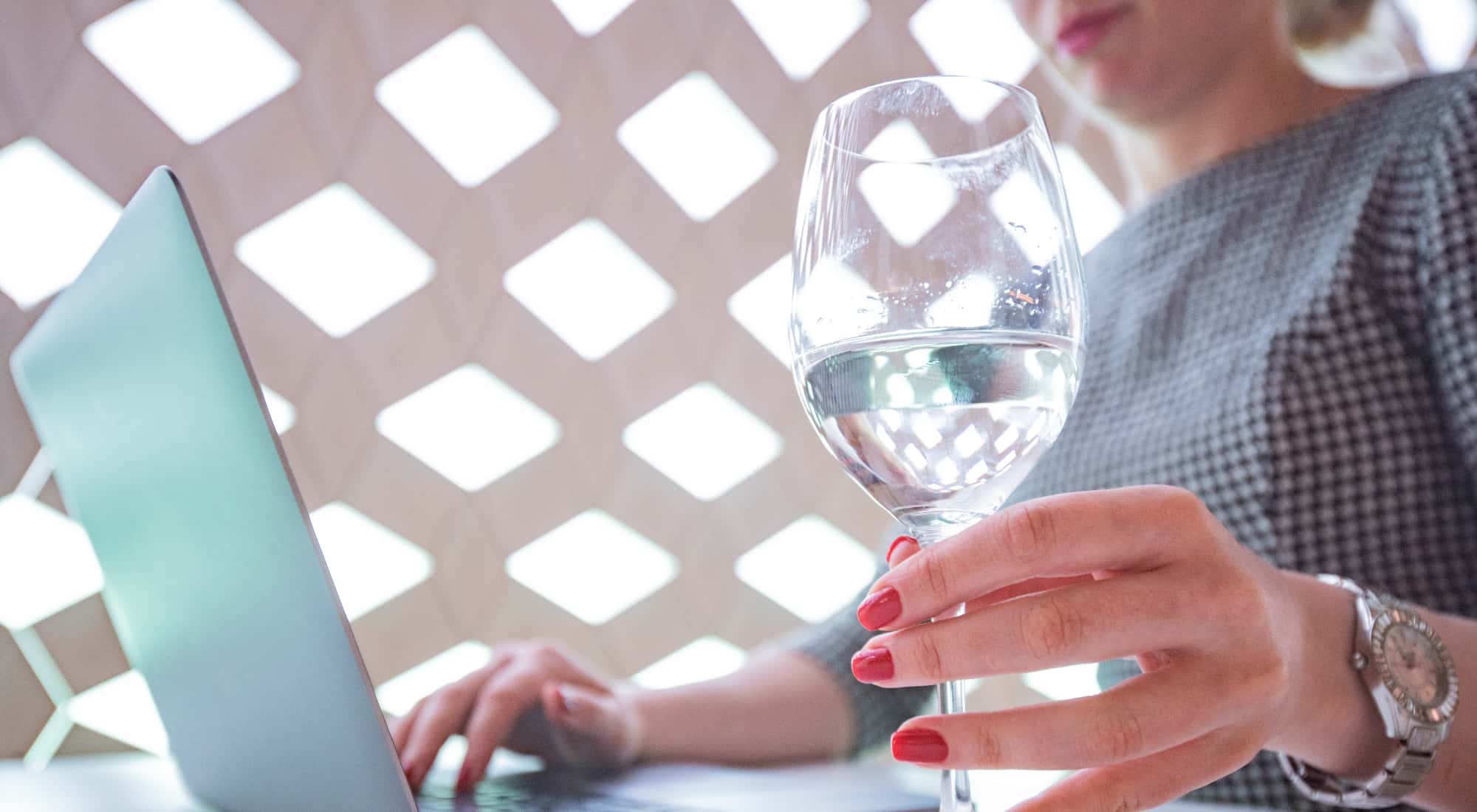 A woman is working on her laptop, and she is holding a wine glass in her hand that has clear, pure water in it.