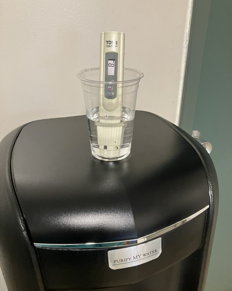 A TDS meter is immmersed in a plastic cup of water that is sitting on top of a black bottleless water cooler. The TDS meter shows a readout of 8 ppm.
